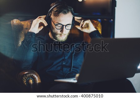Picture bearded man wearing glasses relaxing modern loft office.Banker sitting vintage chair,listening music laptop night.Using contemporary notebook,blurred background.Horizontal,film effect