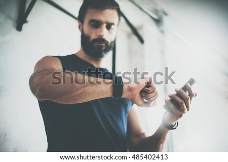 Photo Bearded Sportive Man After Workout Session Checks Fitness Results Smartphone.Adult Guy Wearing Sport Tracker Wristband Arm.Training hard inside gym.Horizontal bar background.Blurred
