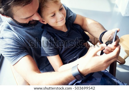 Closeup of young boy sitting with father and using mobile phone in modern sunny place. Horizontal, blurred background