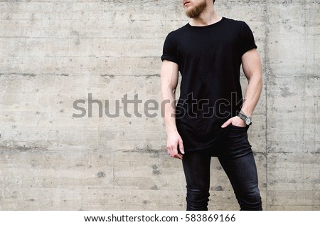 Young muscular bearded man wearing black tshirt and jeans posing in center of modern city. Empty concrete wall on the background. Hotizontal mockup
