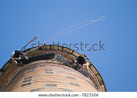 Low angle shot of two unrecognisable workers hanging from a high industrial chimney. Blue sky.