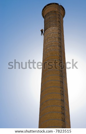Huge Industrial Chimney with unrecognizable worker hanging from it. Blue sky.