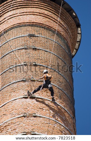 Cropped shot of an unrecognisable worker hanging from a high industrial brick chimney. Blue sky in the background.
