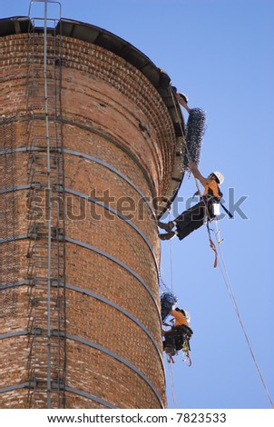 Cropped shot of two unrecognisable workers hanging from a high industrial brick chimney. Blue sky in the background.