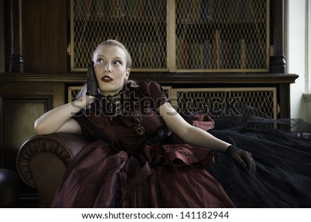 Beautiful Caucasian woman in red Victorian dress lost in thoughts in old library