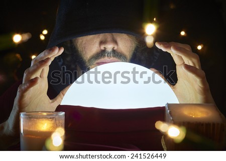 Very low key portrait of hooded man with eyes covered reading fortune on bright crystal ball, surrounded by candle light.