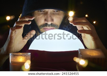 Very low key portrait of hooded man reading fortune on bright crystal ball, surrounded by candle light.