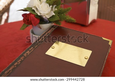 Detail of leather bound menu with blank name plate on red table cloth. There's a small flower vase on the side with red and white roses.
