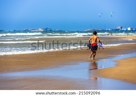 A man in swimming shorts runing along the beach holding the rest of his clothing in his hands
