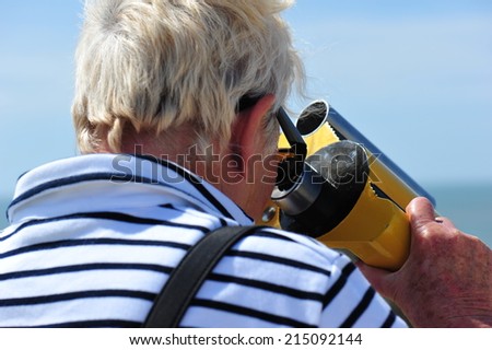 A closeup of an old man is striped shirt looking through the telescope at the sea