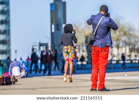 A young man in bright red trousers taking a photo with a lady in colorful shorts walking in front of him