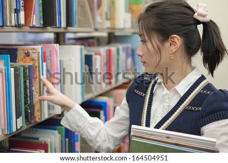 Cute young woman holding books and looking for books on a library shelf