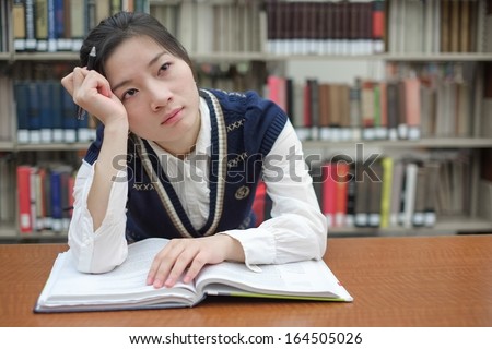 Young girl student with open textbook in front of a library bookshelf looking deep in thought