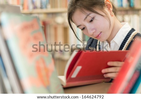 Beautiful female student reading a red covered book on a bookshelf in library