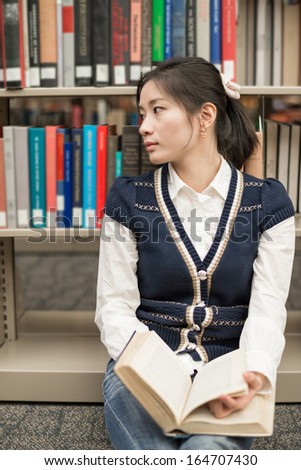 Stressed female student sitting on the floor in front of a bookshelf holding a open thick old book