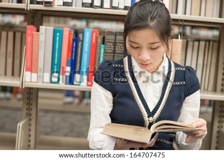 Attractive young girl sitting on the floor in front of a bookshelf reading a thick old book