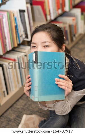 Playful student covering mouth with a blue hard cover book in library
