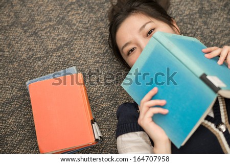 Fun young student laying on carpet with a book covering mouth next to a stack of books
