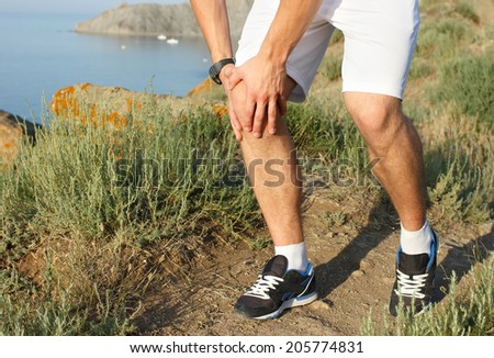 Runner holding sore leg, knee pain from running or exercising, jogging injury or cramp, cross country in summer nature. Caucasian male with tendon ache injured.
