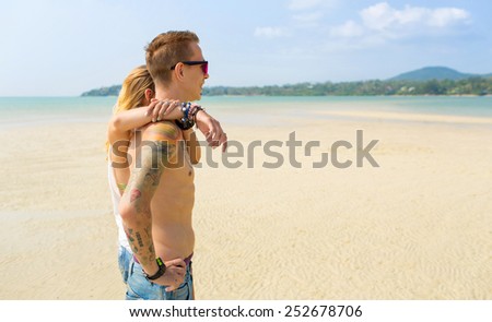 A loving couple standing on the beach side-faced, embracing, enjoying a sunny day in the tropics and looking into the distance