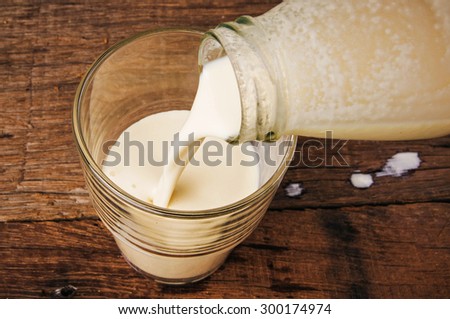 Pouring Fresh Banana Milk from Glasses Bottle into Glass, Ready to Drink, Organic Homemade Dairy Produce. Wood Table Background, Country Rustic Still Life Style.