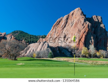 Golf green with flagstick and ball with a dramatic rocky background and blue sky