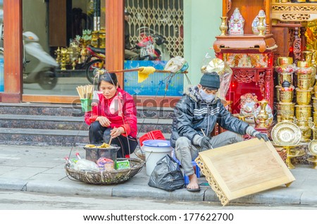 HANOI, VIETNAM, JANUARY 13, 2013 - an artisan is painting a carved table sitting on street while a woman is cooking some food for sale