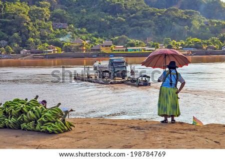 RURRENABAQUE, BOLIVIA - MAY 10, 2014: Local woman in traditional attire looks at ferry on Beni river, transporting a lorry from San Buenaventura on other riverbank