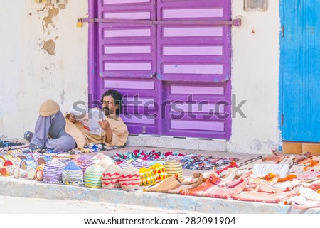 AS-SAWIRA, MOROCCO, APRIL 7, 2015: Street seller of handmade woolen caps waits for customers reading a newspaper