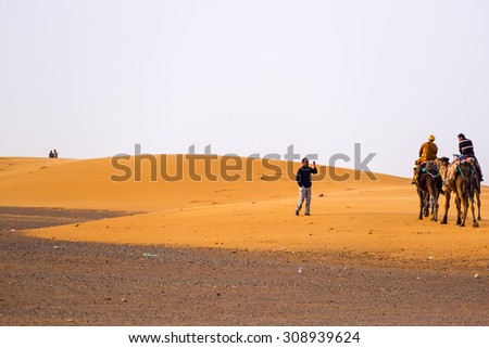 MERZOUGA, MOROCCO, APRIL 13, 2015: Tourist takes photos of people on camels on sand dunes of Erg Chebbi