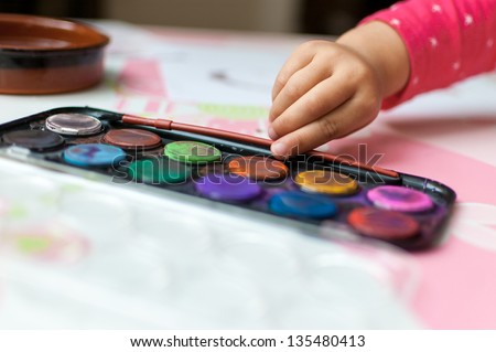 Used watercolor paint tray lies open on table. A little girl is about to pick up her paintbrush.