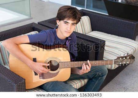 Teenage Boy plays an acoustic guitar while relaxing on a patio chair.