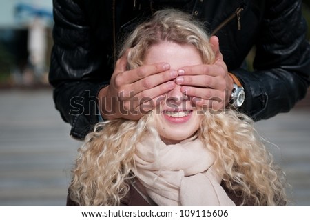 Hands of man covering eyes of young smiling blond woman - outdoor