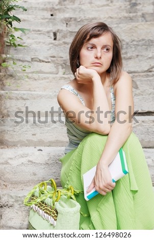 Young woman dressed in green with thoughtful look sits on stairs and holds book