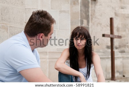 Young couple sitting outdoors ahead of church (cross in background) having relationship problems