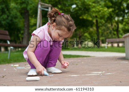 Small cute girl drawing with chalk on sidewalk, trees in background