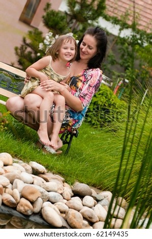 Mother with daughter siting on bench in garden - family house in background