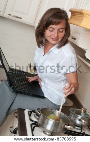 Young woman cooking meal in kitchen according to receipt from laptop (internet)
