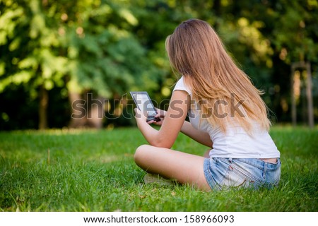 Young woman (teen girl) reading book on electronic book reader - outdoor, rear view