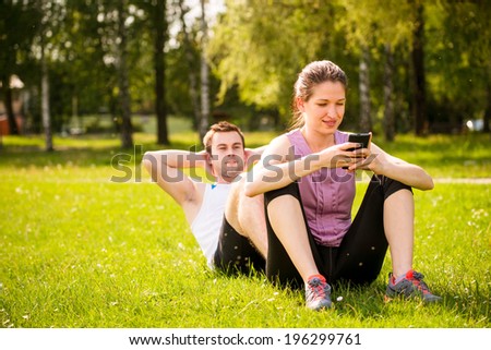 Man making sit-ups while woman is sitting on his feet and playing with mobile phone