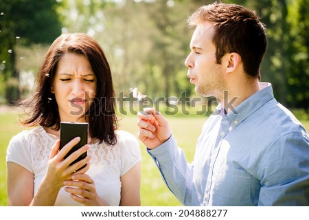 Young man blowing dandelion to face of angry woman holding mobile phone