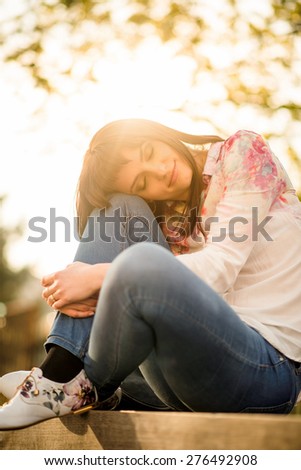Portrait of woman leaning on her knee with closed eyes sitting on bench at sunset