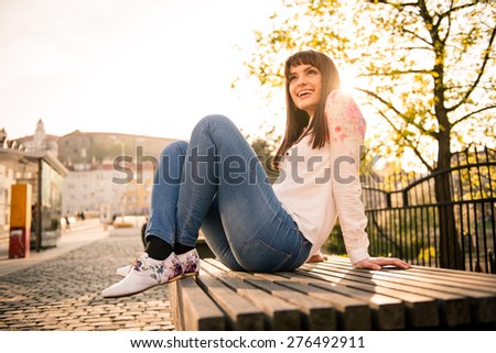 Young woman sitting on bench in street and enjoying life with sun in background