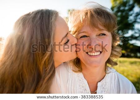 Teenage daughter kissing her mother outdoor in nature with sun in background, wide angle