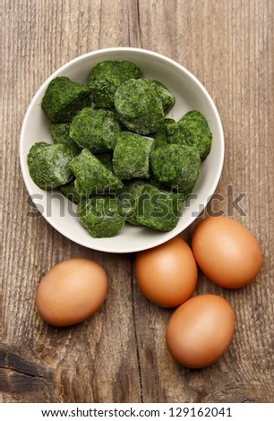 Bowl of frozen spinach isolated on brown wooden background, surrounded by eggs