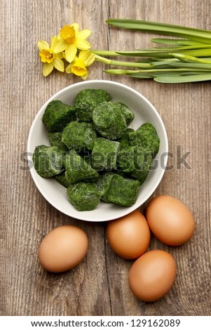 Bowl of frozen spinach isolated on brown wooden background, surrounded by eggs and yellow daffodils