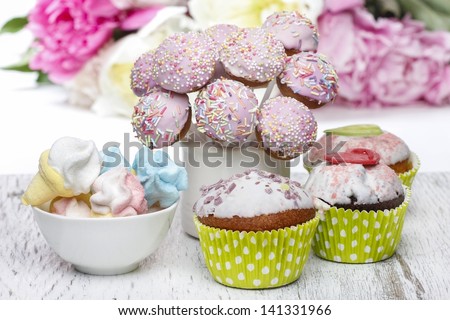 Pastel cake pops, cupcakes and marshmallows on rustic wooden table. Stunning peony flowers in the background.