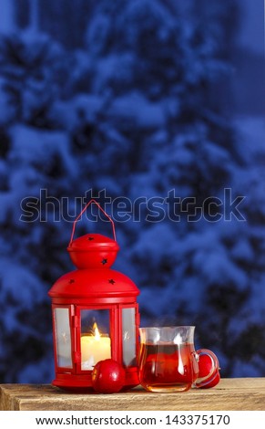 Red lantern, steaming cup of tea and red apples on wooden table. Snowy evening in the background. Copy space.