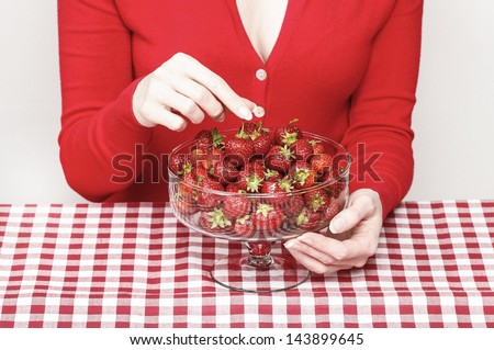 Woman eating strawberries, sitting by the table covered with checkered red and white table cloth, wearing red blouse.