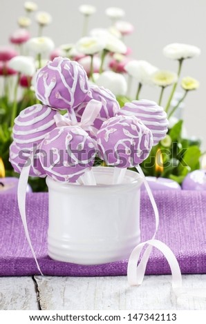 Lilac cake pops in white ceramic jar. White and pink daisies in the background. Party table setting.